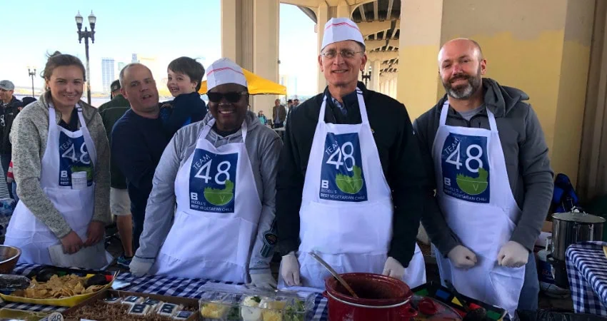 Four people wearing aprons stand in front of a table with food at the Staff Chili Cook Off