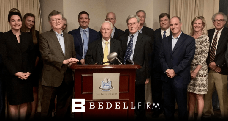 Bedell firm attorneys stand with Charles P. Pillans, III in front of a podium. A white Bedell Firm logo is at the bottom